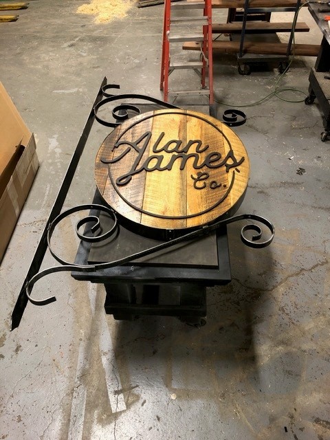 Hand-forged scrolled iron bracket holding a round wood and steel shop sign